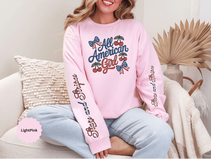 A white crewneck sweatshirt printed with All American Girl in red and blue script with cherries and patriotic bows, with script on both sleeves.