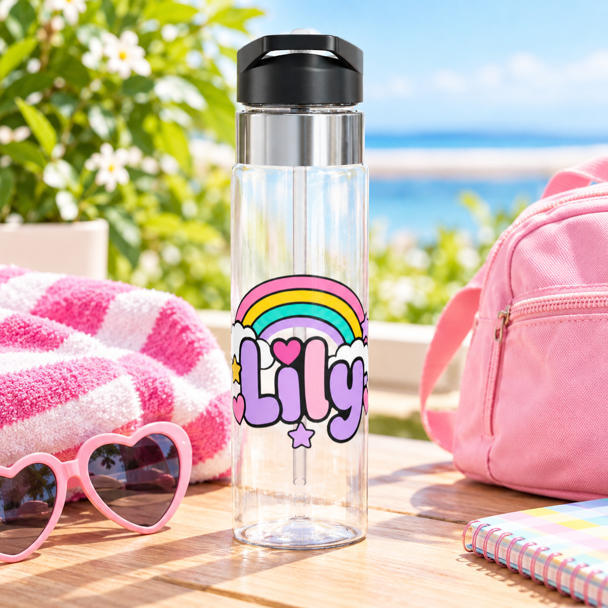 A clear water bottle with a black screw-on lid, stainless band and straw, printed with the sample name "Lily" in lavender letters under a pastel rainbow with white clouds, a pink heart and stars.
