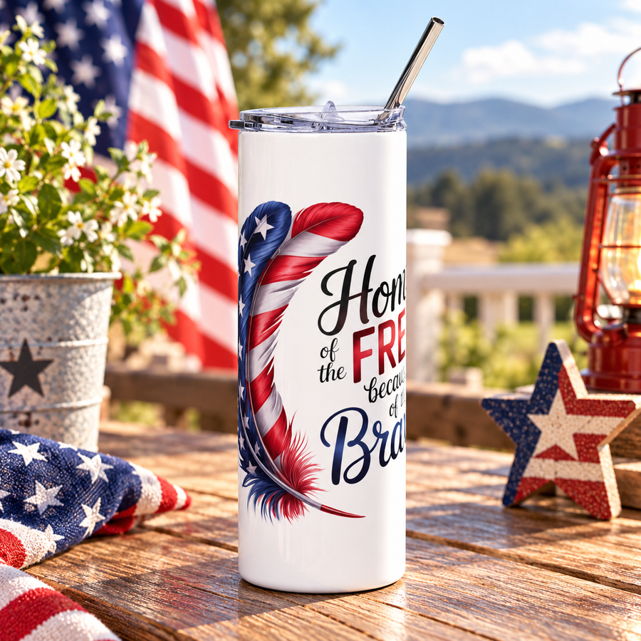 A white stainless steel skinny tumbler with a clear lid and metal straw, printed with a stars-and-stripes feather beside the words "Home of the FREE because of the Brave."