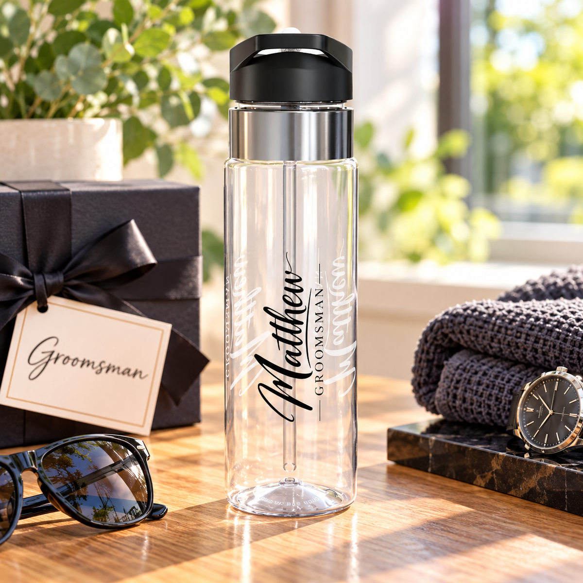 A clear water bottle with a black flip-top lid, stainless band and straw, printed with the sample name "Matthew" in vertical black script above the word "Groomsman".
