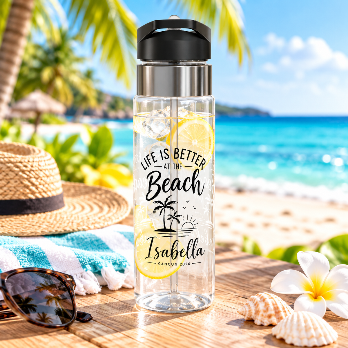 A clear water bottle with a black flip-top lid, stainless band and straw, printed with "Life is better at the Beach" above palm trees and the sample name "Isabella" with "Cancun 2026".