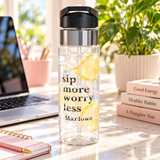 A clear water bottle with a black flip-top lid, stainless band and straw, printed with "sip more worry less" in black lettering above the sample name "Marlowe".