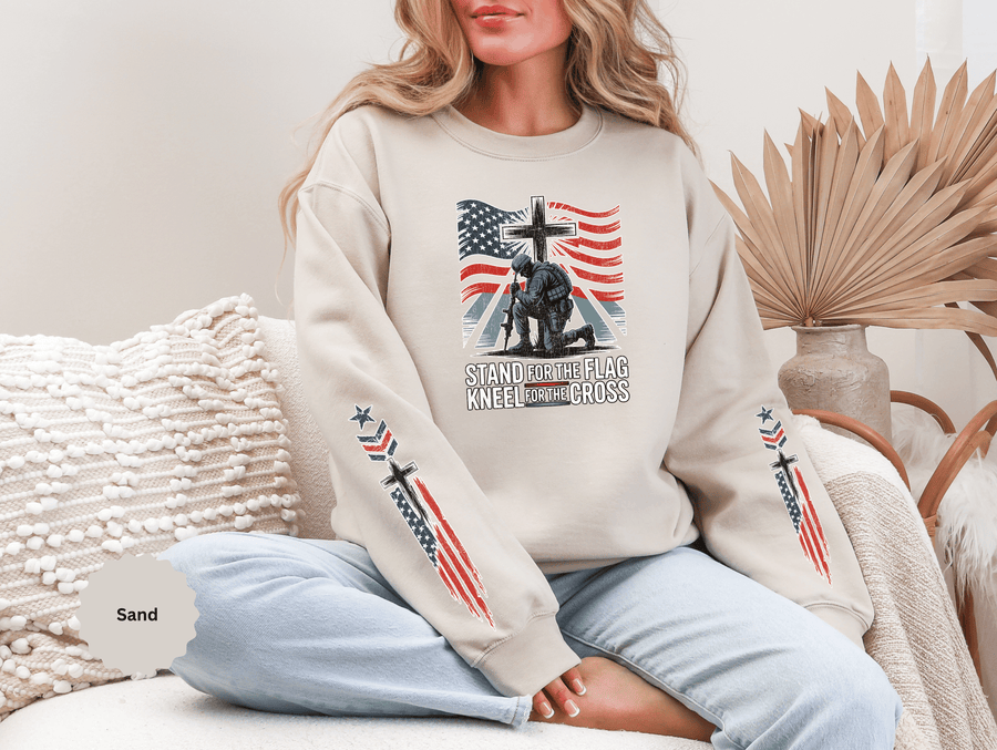 A woman wearing a sand crewneck sweatshirt printed with a soldier kneeling at a cross in front of an American flag above Stand for the Flag, Kneel for the Cross.