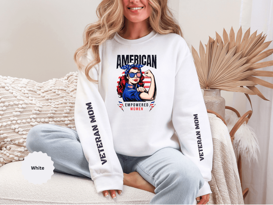 A woman wearing a white crewneck sweatshirt printed with AMERICAN above a woman flexing her arm in a flag bandana and star sunglasses, Empowered Women below, and Veteran Mom down both sleeves.