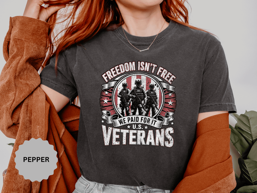 A woman wearing a dark gray t-shirt printed with Freedom Isn't Free above three soldiers on a red flag badge, with We Paid For It and U.S. Veterans below.