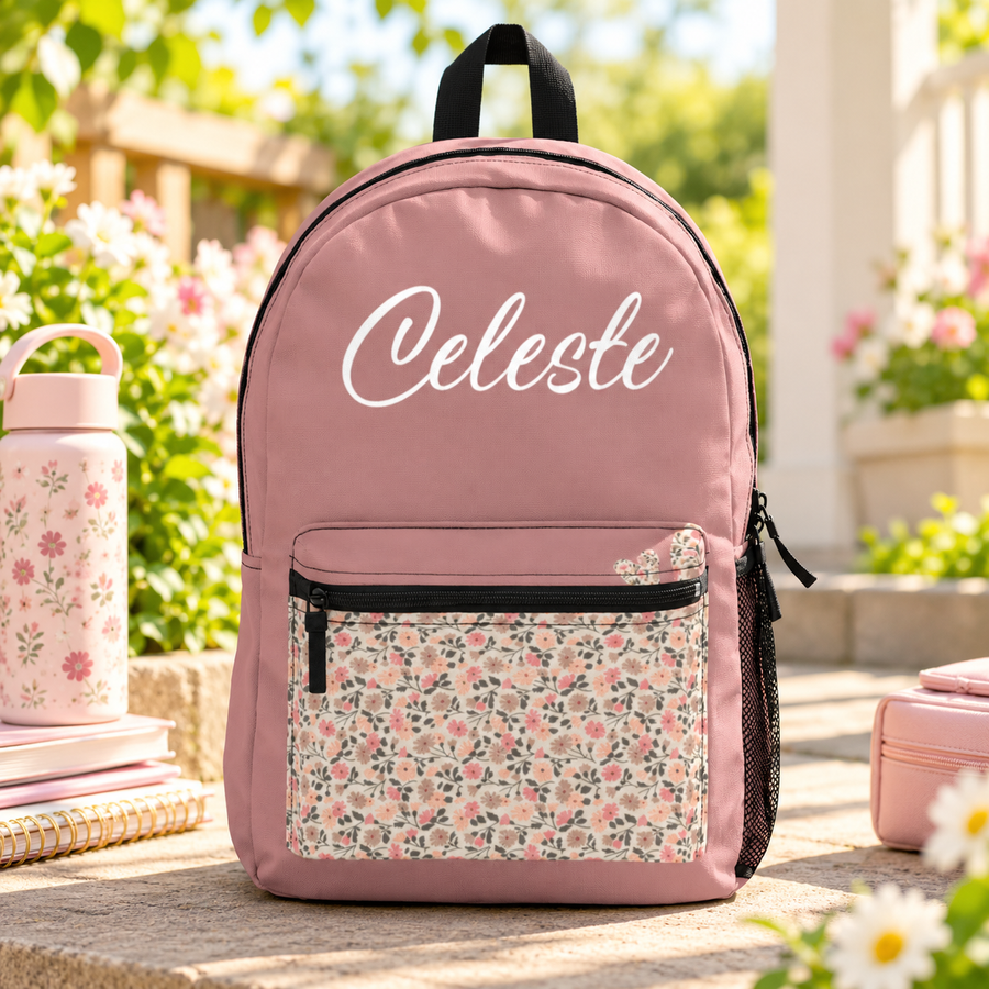 A backpack with a black top handle, front zip pocket and mesh side pocket, dusty pink with a small pink and gray floral print on the front pocket and the sample name "Celeste" in white script across the top.