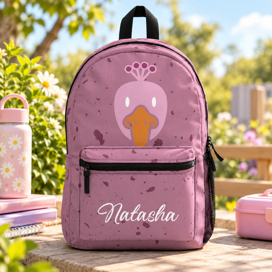 A backpack with a black top handle, front zip pocket and mesh side pocket, speckled pink with a cartoon pink duck face with an orange beak and the sample name "Natasha" in white script on the pocket.