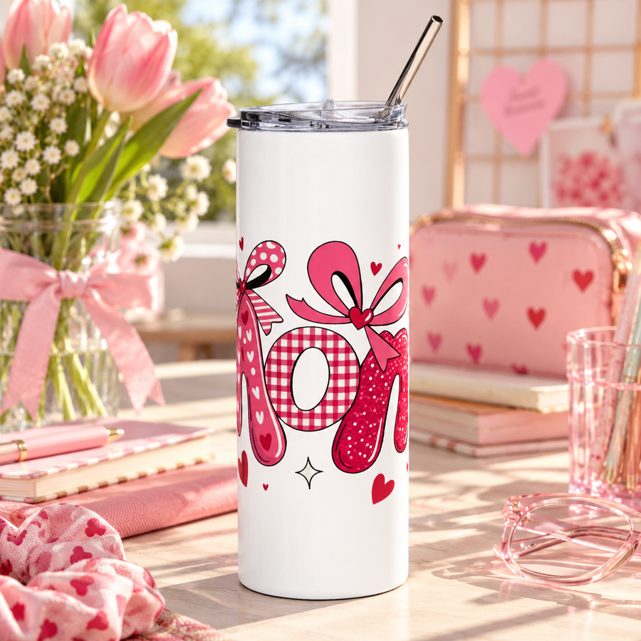 A white stainless steel skinny tumbler with a clear lid and metal straw, printed with "Mom" in pink heart-print, gingham and glitter letters topped with pink bows and small hearts.