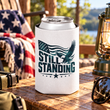A white can cooler on a drink can, printed with "STILL STANDING" in dark green lettering with a soaring bald eagle, a waving American flag and stars.