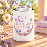 A white can cooler on a drink can, printed with "CREATE YOUR OWN MAGIC" in lilac and coral lettering under a crescent moon and a star wand, framed by butterflies, ribbons and flowers.