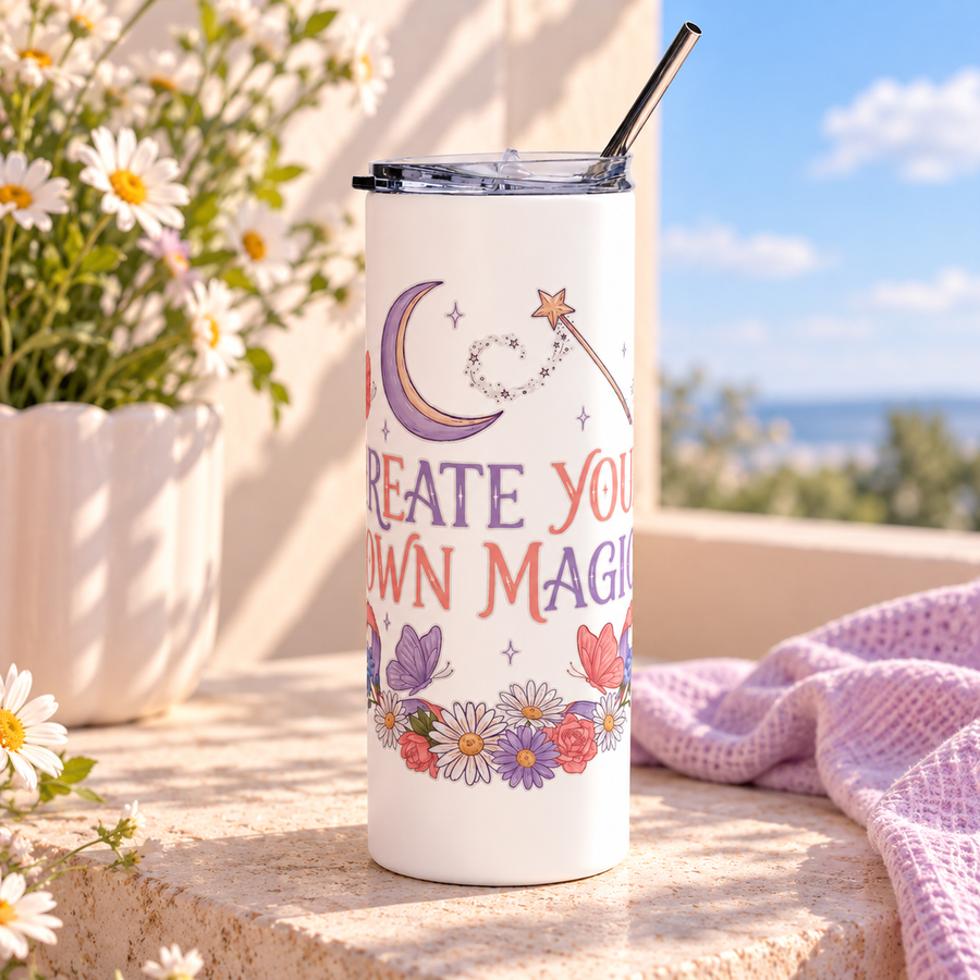 A white stainless steel skinny tumbler with a clear lid and metal straw, printed with "CREATE YOUR OWN MAGIC" in lilac and coral lettering under a crescent moon and a star wand, framed by butterflies, ribbons and flowers.