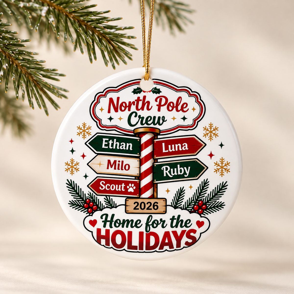 A round white ceramic Christmas ornament on a gold string, printed with "North Pole Crew" above a candy-cane signpost with five sample names, "2026" and "Home for the Holidays".
