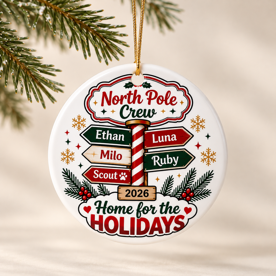 A round white ceramic Christmas ornament on a gold string, printed with "North Pole Crew" above a candy-cane signpost with five sample names, "2026" and "Home for the Holidays".