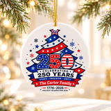 A round white ceramic Christmas ornament on a gold string, printed with a red, white and blue Christmas tree with stars, a large "250", "Celebrating 250 Years", a sample family name and "1776–2026".
