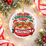 A round white ceramic Christmas ornament on a gold string, printed with "COZY CHRISTMAS CREW" above five gingerbread people in Santa hats, each with a sample name, in a red mug reading "The Parker Family, Baked with Love 2026".