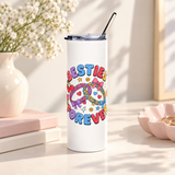 A white stainless steel skinny tumbler with a clear lid and metal straw, printed with "BESTIES FOREVER" in colorful bubble lettering around two linked braided friendship bracelets with bows, hearts, cherries, stars and smiley faces.