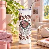 A white insulated tumbler with a clear lid and metal straw sits on a wooden table in a bright, pink-themed room. The tumbler is printed with brown script text reading 'Always Moving' and an illustration of pink flowers and white sneakers.