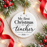 A round white ceramic Christmas ornament on a gold string, printed with "My first Christmas as a teacher" in black serif and script lettering, with "A milestone to remember" and "2026".