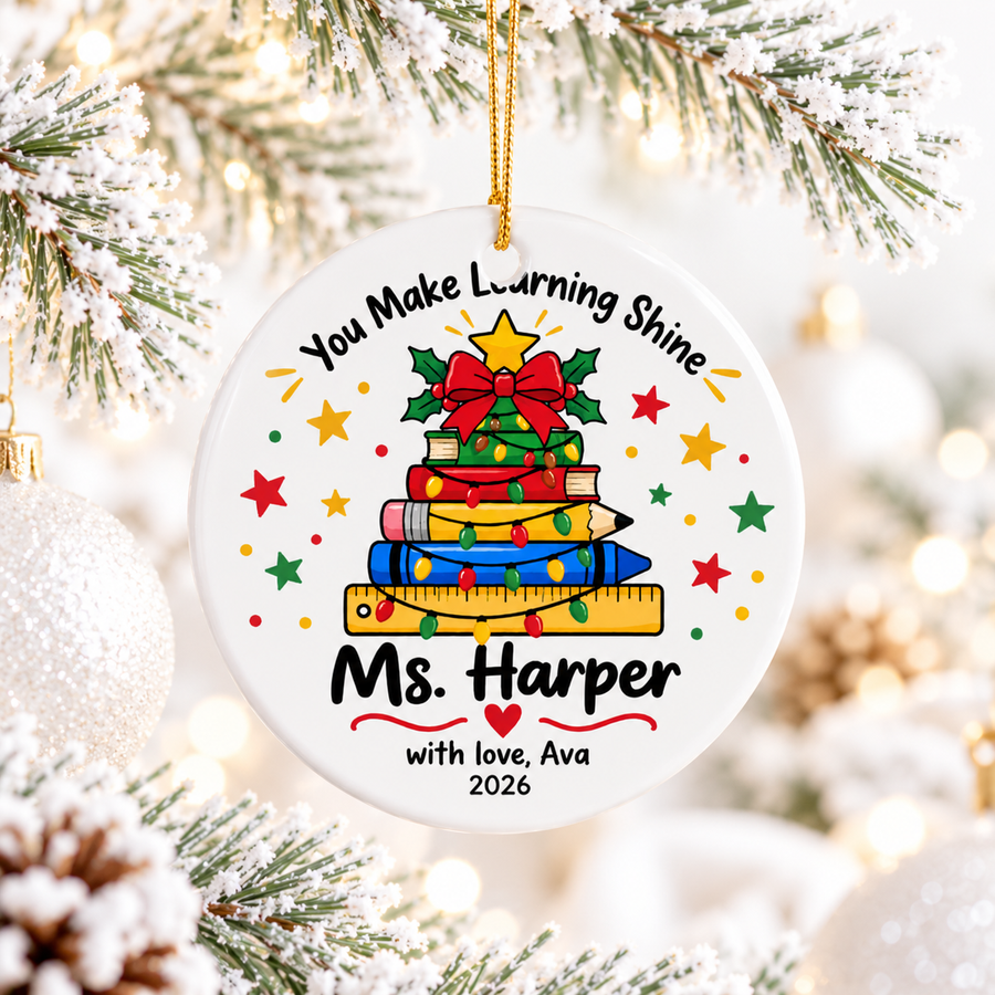 A round white ceramic Christmas ornament on a gold string, printed with "You Make Learning Shine" above a Christmas tree made of stacked books, a pencil and a ruler wrapped in lights, with a sample teacher name, "with love" signature and "2026".