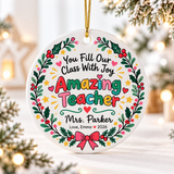 A round white ceramic Christmas ornament on a gold string, printed with "You Fill Our Class With Joy" and "Amazing Teacher" in colorful lettering inside a holly and berry wreath with a pink bow, with a sample teacher name and a "Love" signature with "2026".