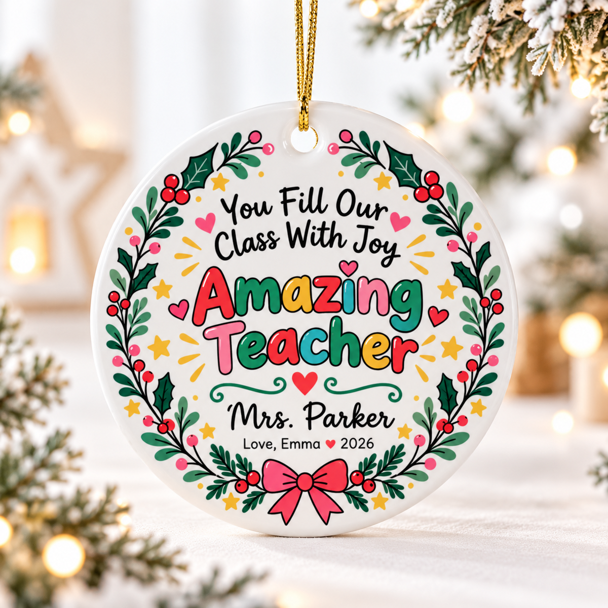A round white ceramic Christmas ornament on a gold string, printed with "You Fill Our Class With Joy" and "Amazing Teacher" in colorful lettering inside a holly and berry wreath with a pink bow, with a sample teacher name and a "Love" signature with "2026".