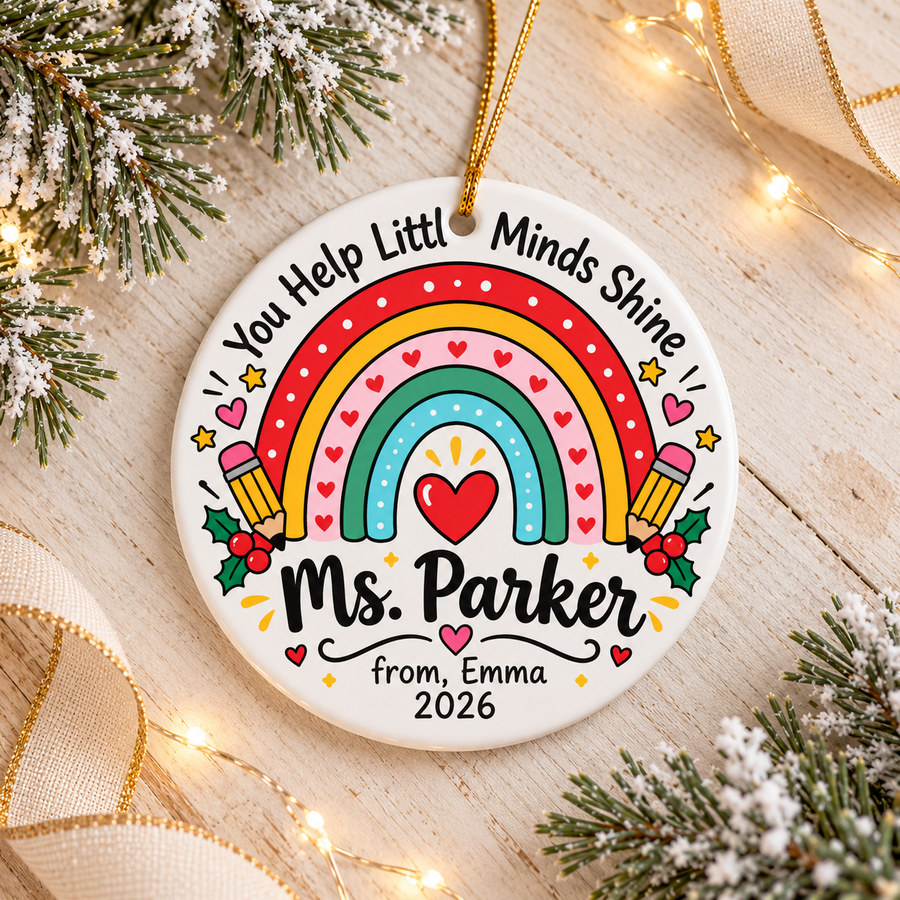 A round white ceramic Christmas ornament on a gold string, printed with a red, yellow, pink, green and blue rainbow with a red heart, two pencils and holly under arched "You Help Little Minds Shine" lettering, with a sample teacher name, a "from" signature and "2026".