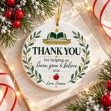 A round white ceramic Christmas ornament on a gold string, printed with an open green book under a gold star on a banner with a sample school name, "THANK YOU for helping us learn, grow & believe", "2026", an apple and a "Love" signature, framed by a leafy wreath.