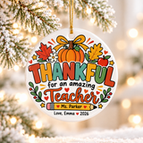 A round white ceramic Christmas ornament on a gold string, printed with "THANKFUL for an amazing Teacher" in colorful lettering under a pumpkin with a yellow bow, autumn leaves and an apple, with a sample teacher name on a pencil and a "Love" signature with "2026".
