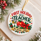 A round white ceramic Christmas ornament on a gold string, printed with "FIRST HOLIDAY AS A TEACHER" in red and green lettering above a red apple, a cup of pencils, stacked books with a red bow and holly, and a banner with a sample teacher name and "2026".