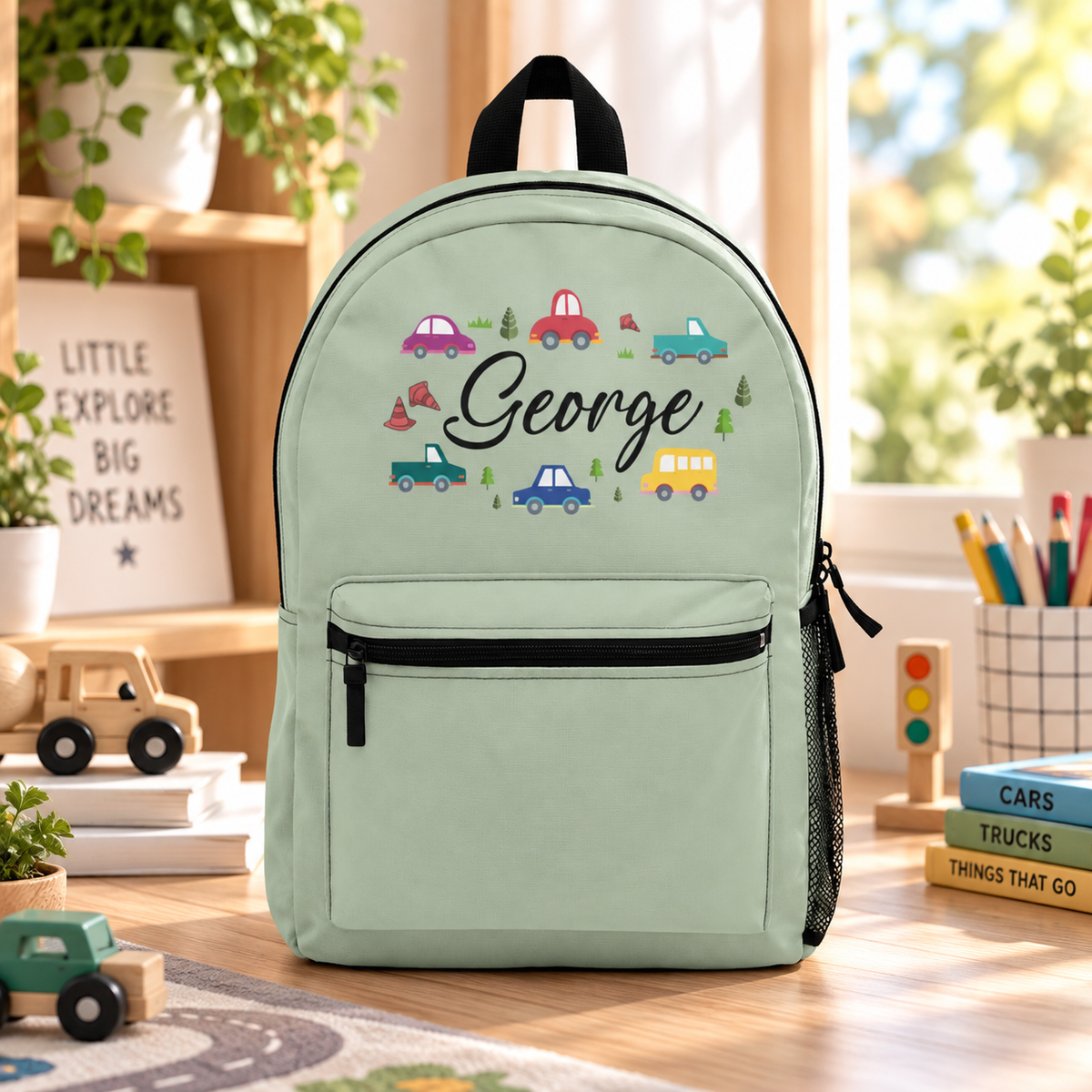 A kids backpack with a black top handle, front zip pocket and mesh side pocket, sage green with small colorful cars, a truck, a bus, traffic cones and trees around the sample name "George" in black script.