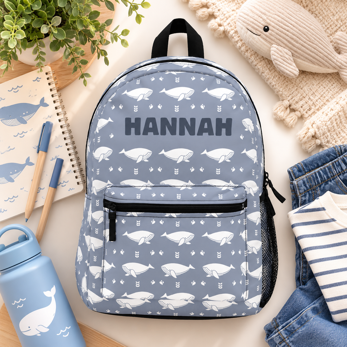A kids backpack with a black top handle, front zip pocket and mesh side pocket, blue gray with an all-over pattern of white whales and small arrows and the sample name "HANNAH" in bold dark lettering.