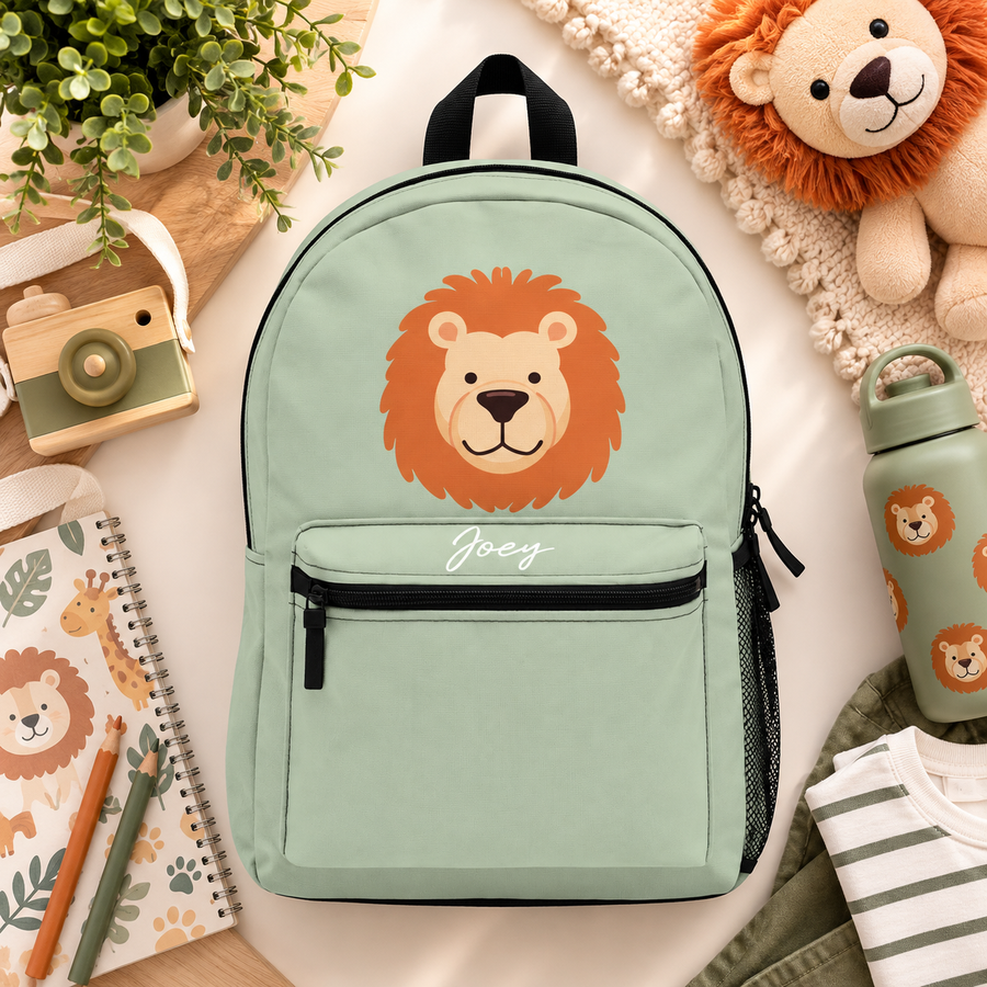 A kids backpack with a black top handle, front zip pocket and mesh side pocket, sage green with a large friendly lion face with an orange mane and the sample name "Joey" in white script on the pocket.