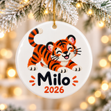 A round white ceramic Christmas ornament on a gold string, printed with a smiling orange and black striped tiger cub in an embroidery style, above a sample child's name in black and "2026" in orange.