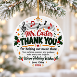A round white ceramic Christmas ornament on a gold string, printed with a treble clef and red, green and black notes on a staff above a sample teacher name, "THANK YOU for helping our music shine", a short note of thanks, holly, "Warm Holiday Wishes" and a "Love" signature with "2026".