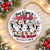 A round white ceramic Christmas ornament on a gold string, printed with "Our Class is WILD about" a sample teacher name in black and pink lettering around three cartoon white geese honking, with speech bubbles reading "Honk!", "Best Teacher!" and a cheer, hearts, stars and a room-number signature with "2026".