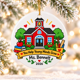 A round white ceramic Christmas ornament on a gold string, printed with a red schoolhouse with a bell tower between green trees, an apple, stacked books and a cup of pencils and a ruler, above a yellow banner reading "You Help Young Minds Grow", a sample teacher name and "2026".