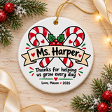 A round white ceramic Christmas ornament on a gold string, printed with two crossed red and white candy canes with holly and berries behind a banner with a sample teacher name, "Thanks for helping us grow every day", hearts and a "Love" signature with "2026".