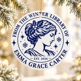 A round white ceramic Christmas ornament on a gold string, printed with "FROM THE WINTER LIBRARY OF" and a sample name in navy serif lettering around a classic portrait of a woman with holly in her hair, snowflakes, stars and "EST. 2026".