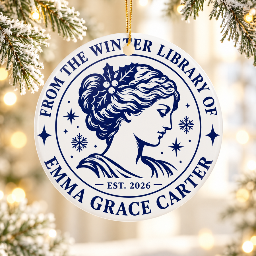A round white ceramic Christmas ornament on a gold string, printed with "FROM THE WINTER LIBRARY OF" and a sample name in navy serif lettering around a classic portrait of a woman with holly in her hair, snowflakes, stars and "EST. 2026".