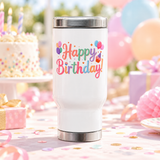 A white stainless steel travel mug with a silver lid and base, printed with "Happy Birthday!" in rainbow pastel lettering with bunches of balloons, a cupcake and small hearts.