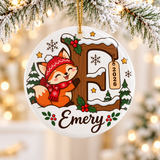 A round white ceramic Christmas ornament on a gold string, printed with a smiling fox in a red knit hat and scarf hugging a large snowy wooden letter with holly and a "2026" tag, among pine trees and snowflakes, above a sample child's name in brown script.