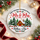 A round white ceramic Christmas ornament on a gold string, printed with "FIRST HOLIDAY AS NEWLYWEDS" arched over "Mr. & Mrs." in red script, a snowy log cabin among pine trees with a fence, sample couple names and a sample wedding date.