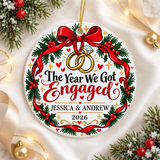 A round white ceramic Christmas ornament on a gold string, printed with two linked gold rings, one with a diamond, hanging from a red bow on a holly wreath, around "The Year We Got Engaged" in black and red lettering, sample couple names and "2026".