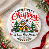 A round white ceramic Christmas ornament on a gold string, printed with "OUR FIRST Christmas" in black and red lettering over a snowy cottage with glowing windows, a wreath on the door, pine trees and a fence, then "in Our New Home", sample couple names and "2026".