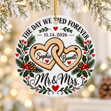 A round white ceramic Christmas ornament on a gold string, printed with "THE DAY WE TIED FOREVER" arched over a sample wedding date, two linked wooden-look hearts with sample names, holly, pinecones and red berries, and "Mr. & Mrs." in black script.