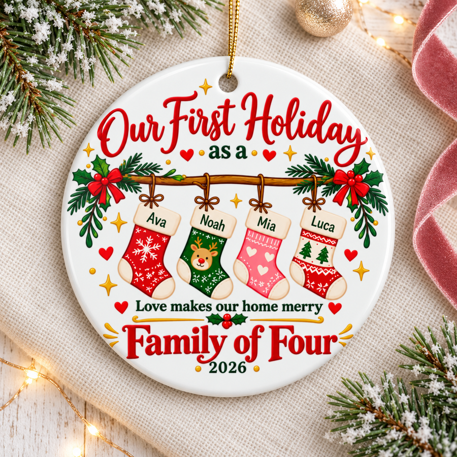 A round white ceramic Christmas ornament on a gold string, printed with "Our First Holiday as a" in red script over four Christmas stockings hung from a holly garland, each with a sample name, then "Love makes our home merry", "Family of Four" and "2026".