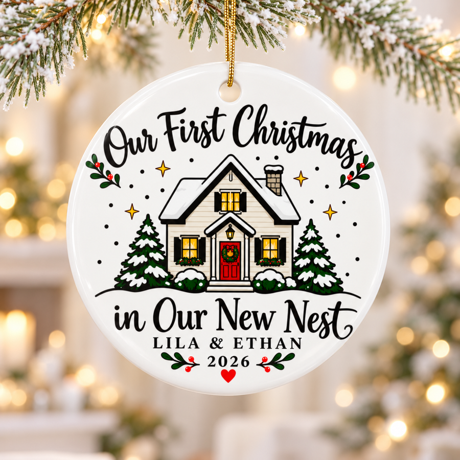A round white ceramic Christmas ornament on a gold string, printed with "Our First Christmas in Our New Nest" in black script around a snow-covered white house with a red door, glowing windows and snowy pine trees, with sample couple names, "2026" and a red heart.