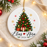 A round white ceramic Christmas ornament on a gold string, printed with a decorated Christmas tree with red and gold baubles, bows and wrapped presents under a gold star, above two sample names in black script joined by a red heart and "2026".