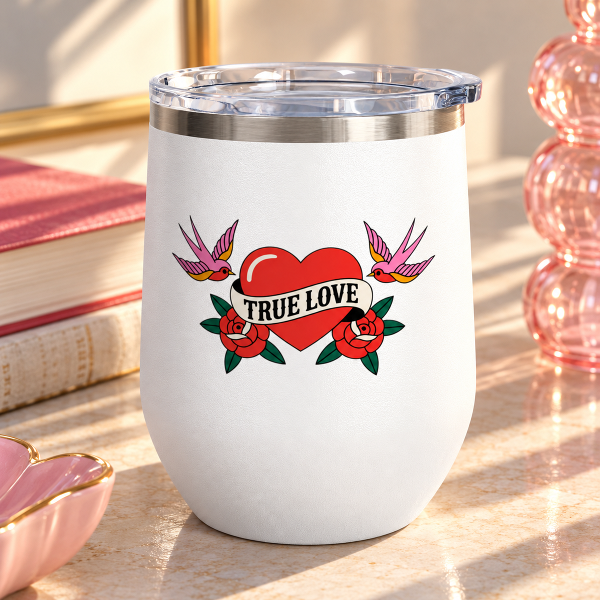 A white stainless steel stemless wine tumbler with a clear push-on lid, printed with a red heart wrapped in a "TRUE LOVE" banner between two red roses, with a pink swallow on each side, in retro tattoo style.