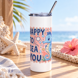 A white stainless steel skinny tumbler with a clear lid and metal straw, printed with "HAPPY TO SEA YOU" in navy lettering beside a smiling blue seahorse with bubbles, coral and a shell on a peach background.